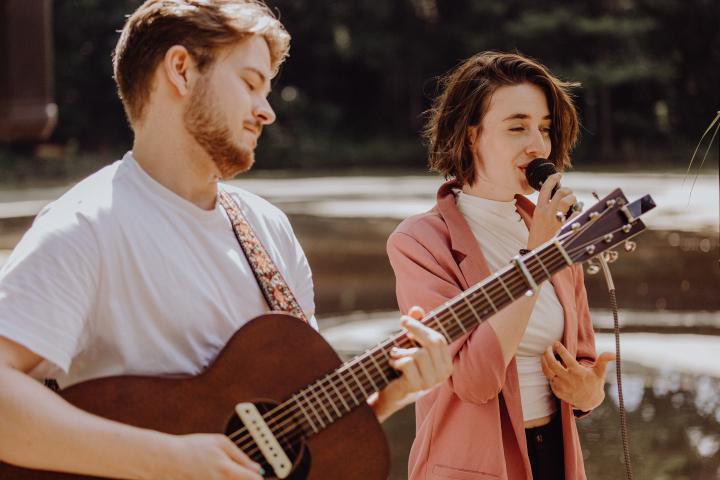 Ein Mann spielt Gitarre und eine Frau singt mit Mikrofon im Freien vor einem unscharfen Hintergrund mit Wasser und Bäumen.