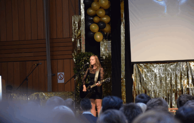 Junge Frau in schwarzem Kleid mit Spitzenärmeln spielt auf einer Bühne Gitarre und singt vor Publikum, im Hintergrund goldene und schwarze Ballons sowie goldene Dekorationen.