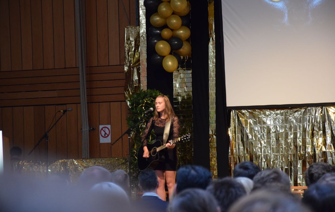 Junge Frau in schwarzem Kleid mit Spitzenärmeln spielt auf einer Bühne Gitarre und singt vor Publikum, im Hintergrund goldene und schwarze Ballons sowie goldene Dekorationen.