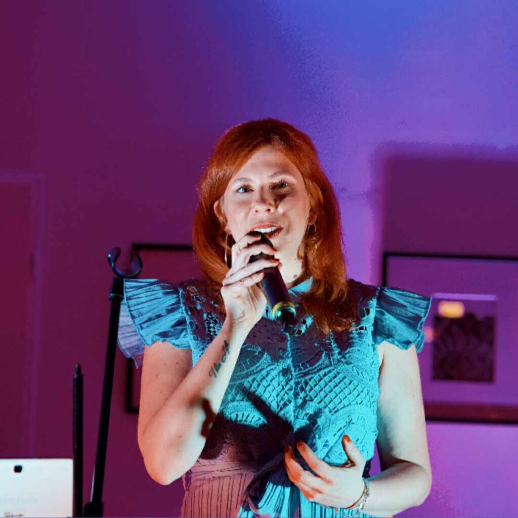 Frau mit roten Haaren und blauem, mit Spitzen verzierendem Oberteil hält ein Mikrofon und spricht vor einem lila beleuchteten Hintergrund.