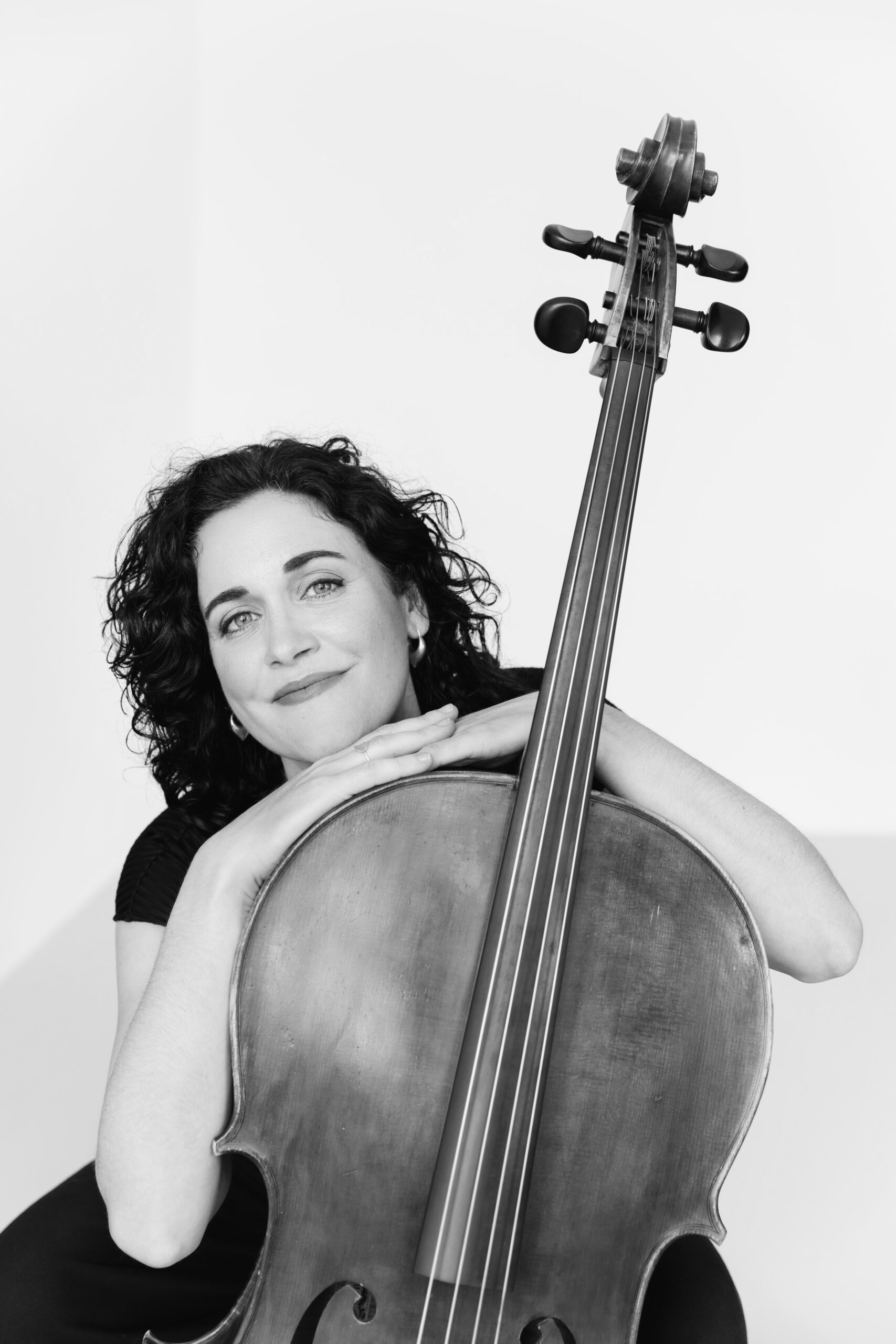 Schwarz-weiß-Porträt einer lächelnden Frau mit lockigem Haar, die mit verschränkten Armen auf einem Cello lehnt.