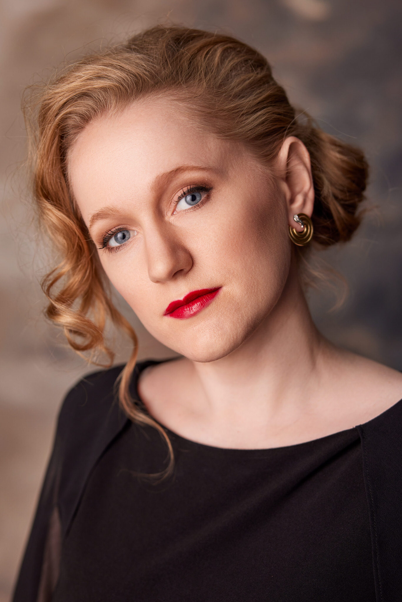 Porträt einer Frau mit blauen Augen, roten Lippen, kupferrotem welligem Haar, hochgesteckt und einem lockeren Strang vorn, sie trägt goldene Kreolenohrringe und ein schwarzes Oberteil, vor unscharfem braun-grauem Hintergrund