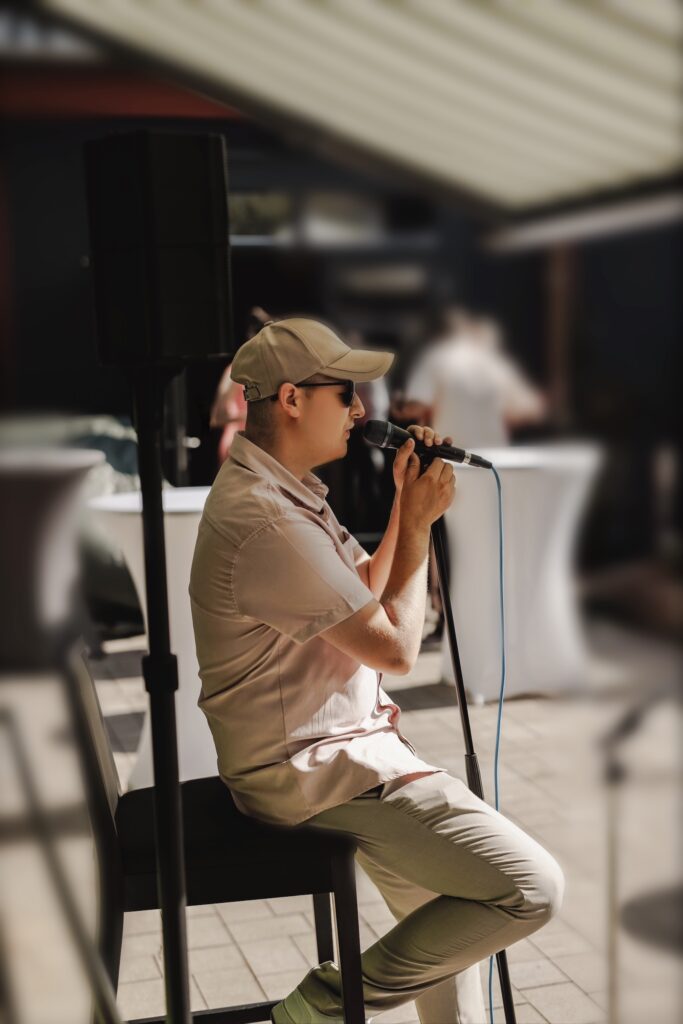 Eine Person sitzt auf einem Stuhl und singt in ein Mikrofon, trägt eine Kappe, Sonnenbrille und helle Kleidung, im Hintergrund unscharfe stehende Tische und Personen.