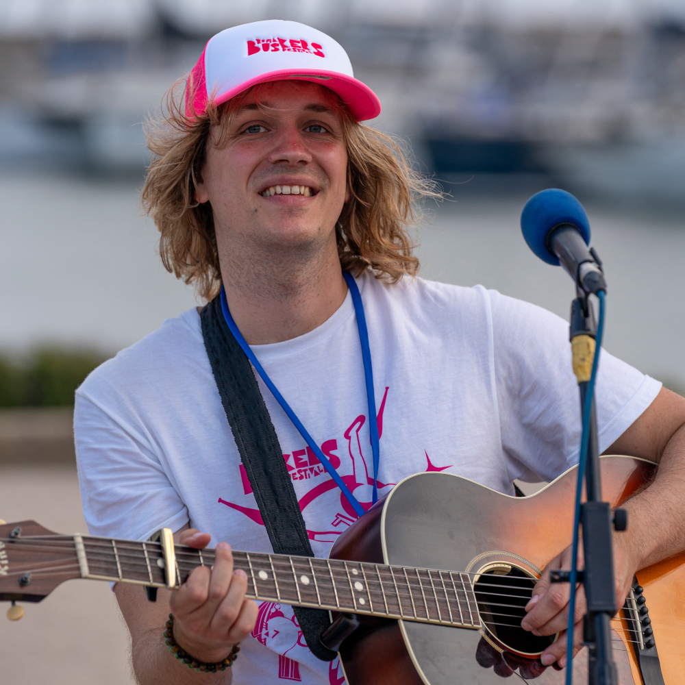Junger Mann mit lockigen blonden Haaren, weißem T-Shirt und rotem-weißem Baseballcap spielt Gitarre vor einem Mikrofon mit blauem Schaumstoffüberzug im Freien, unscharfer Hintergrund mit Wasser und Booten