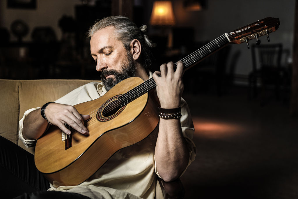 Mittelalterlicher Mann mit grauem Dutt und Bart spielt Akustikgitarre entspannt auf einem Sofa in einem gedämpft beleuchteten Raum.
