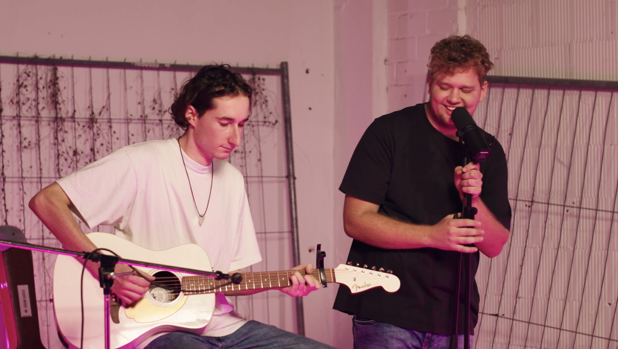 Zwei junge Männer in einem Innenraum mit rosa Beleuchtung, einer spielt Gitarre und der andere singt in ein Mikrofon.