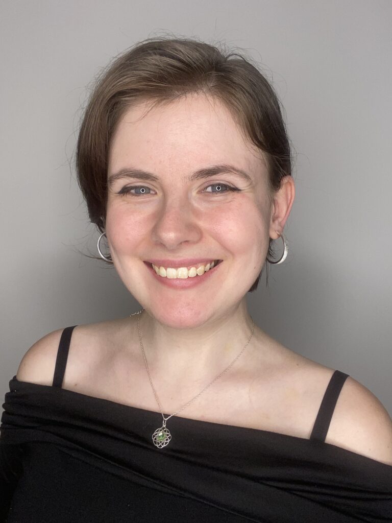 Porträt einer lächelnden jungen Frau mit kurzen braunen Haaren, die silberne Ohrringe und eine Halskette mit einem grünen Anhänger trägt, vor einem neutral grauen Hintergrund.