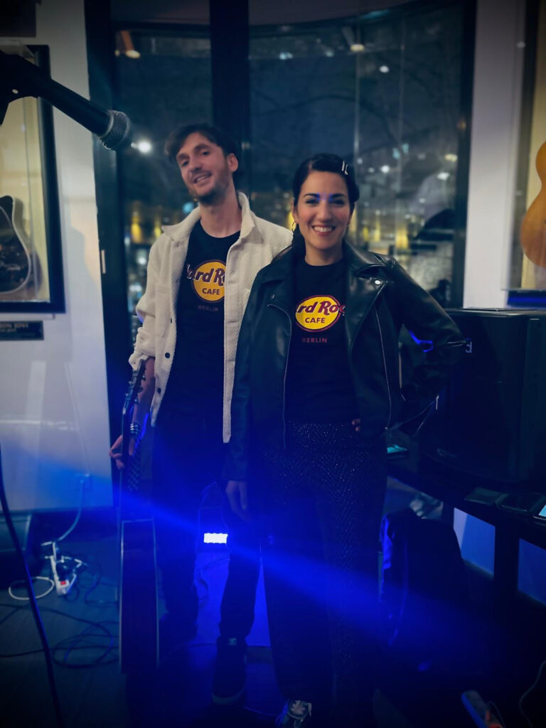 Ein Mann und eine Frau stehen nebeneinander in einem Raum mit dunkler Atmosphäre, beide tragen schwarze Hard Rock Cafe Berlin T-Shirts, der Mann hält eine Gitarre und trägt eine weiße Jacke, die Frau trägt eine schwarze Lederjacke und lächelt.