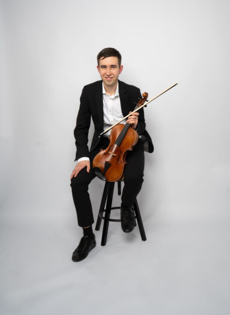 Junger Mann in schwarzem Anzug sitzt auf einem schwarzen Hocker vor weißem Hintergrund und hält eine Violine mit Bogen in den Händen.