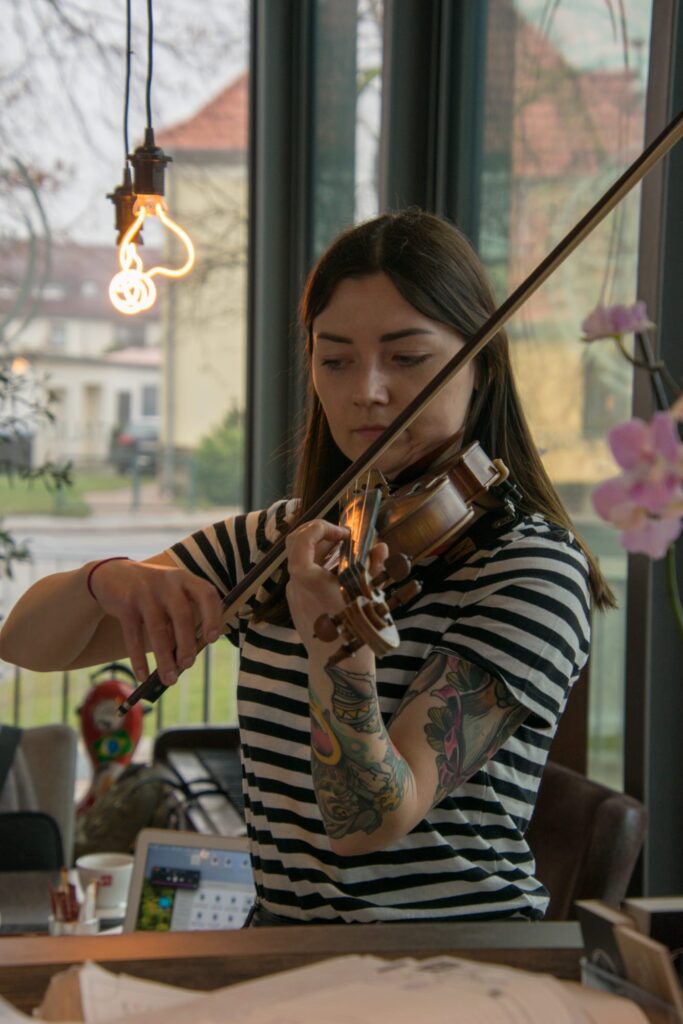 Frau mit langen dunklen Haaren und gestreiftem T-Shirt spielt Violine in einem Raum mit großen Fenstern, sichtbare bunte Tätowierungen auf ihrem rechten Arm, im Hintergrund Computerbildschirm und hängende Lichtquelle