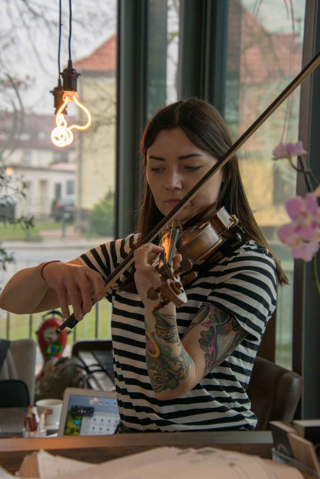 Frau mit langen dunklen Haaren und gestreiftem T-Shirt spielt Violine in einem Raum mit großen Fenstern, sichtbare bunte Tätowierungen auf ihrem rechten Arm, im Hintergrund Computerbildschirm und hängende Lichtquelle