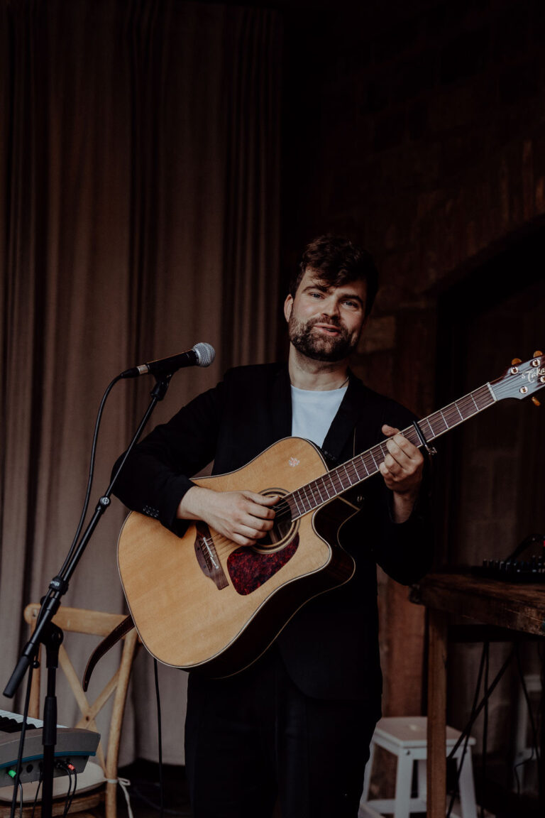 Mann mit dunklem Sakko spielt akustische Gitarre und steht vor einem Mikrofon in einem Raum mit dunklem Hintergrund und Vorhängen.