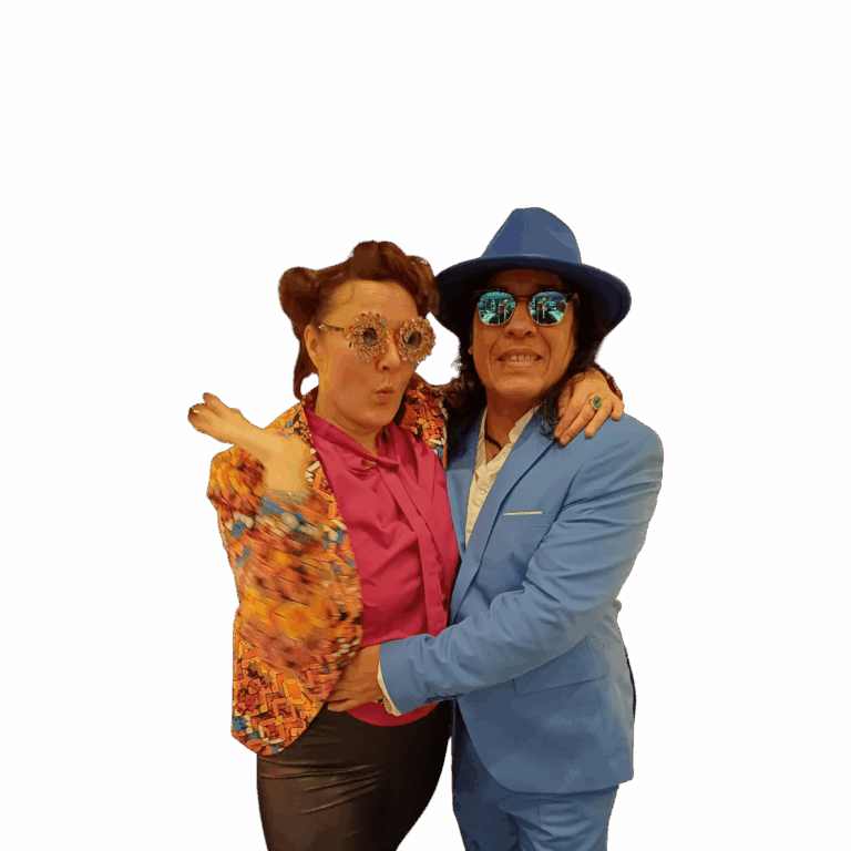 Ein Paar in ausgefallener Kleidung, die Frau trägt eine bunte gemusterte Jacke, eine pinke Bluse und auffällige, mit Juwelen dekorierte Brille, der Mann trägt einen hellblauen Anzug, einen passenden Hut und reflektierende Sonnenbrillen, beide umarmen sich und posieren fröhlich für die Kamera.