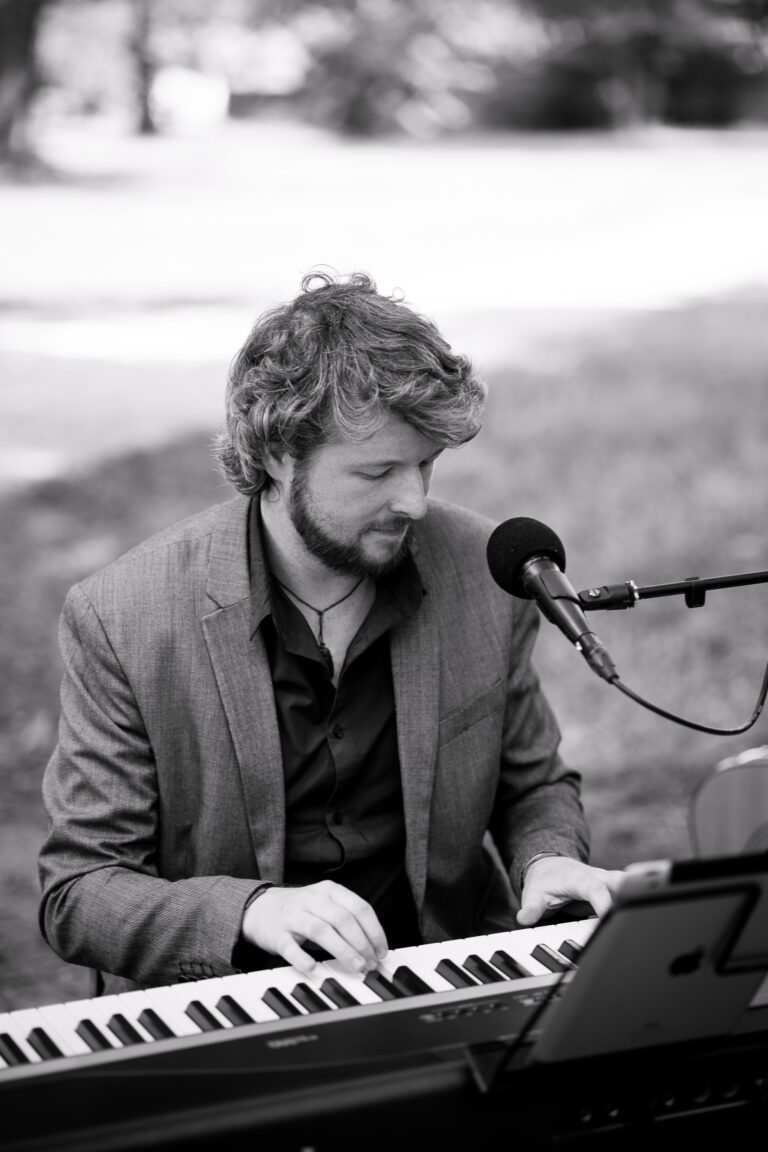 Schwarz-Weiß-Foto eines Mannes mit kurzen lockigen Haaren und Bart, der einen Blazer und ein Hemd trägt und an einem Keyboard spielt, mit einem Mikrofon vor ihm und einem Tablet als Notenständer