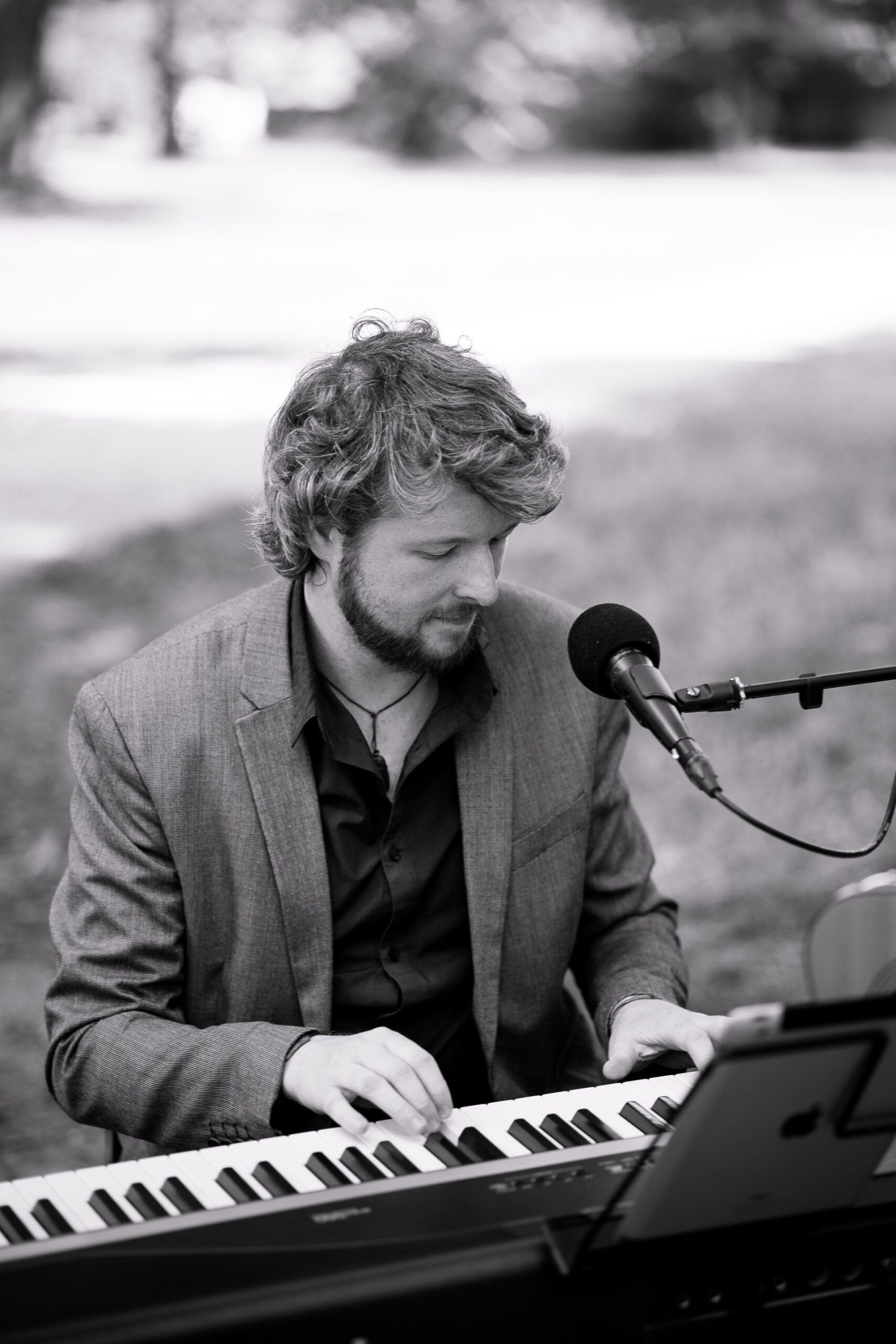 Schwarz-Weiß-Foto eines Mannes mit kurzen lockigen Haaren und Bart, der einen Blazer und ein Hemd trägt und an einem Keyboard spielt, mit einem Mikrofon vor ihm und einem Tablet als Notenständer