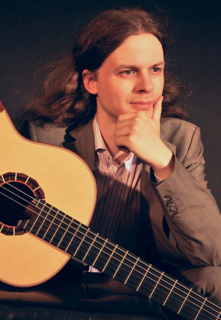 Mann mit langen braunen Haaren in Anzug, der nachdenklich zur Seite schaut und eine Gitarre hält.