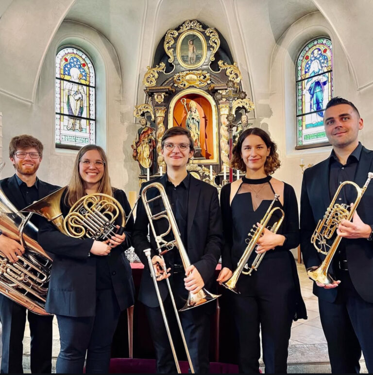 Fünf Musiker in schwarzer Kleidung stehen mit ihren Blechblasinstrumenten in einer Kirche vor einem verzierten Altar mit Statuen und bunten Glasfenstern.