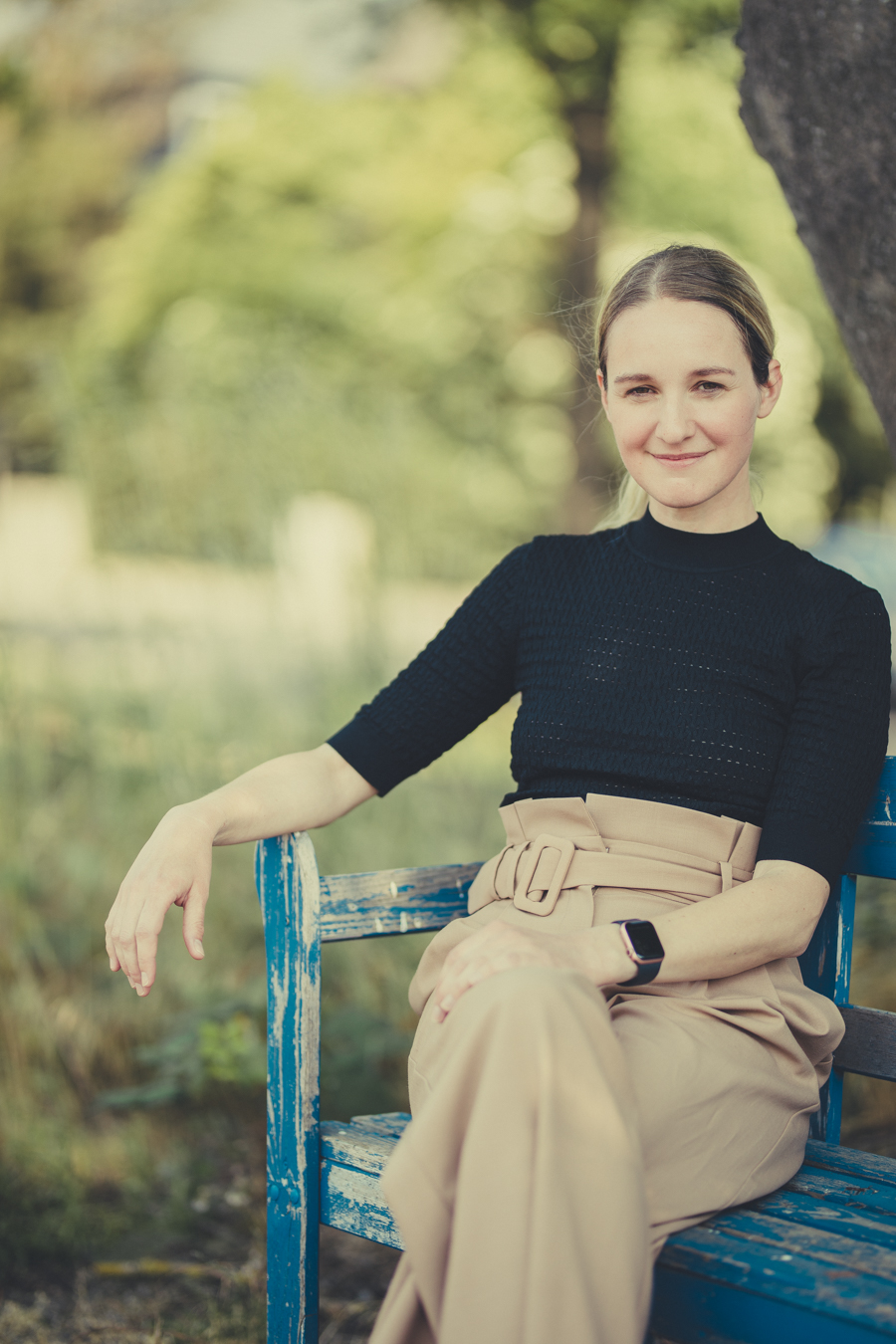 Stefanie Mai mit zurückgebundenem Haar, die auf einer blauen Bank im Freien sitzt und lächelt, trägt schwarzen Pullover und beige Hose mit Gürtel, umgeben von unscharfem grünem Hintergrund.