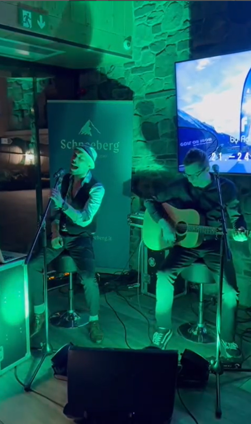 Zwei Musiker sitzen auf Barhockern vor einer Steinwand mit grünem Licht, einer singt in ein Mikrofon, der andere spielt Gitarre, im Hintergrund ein Bildschirm und ein Banner mit der Aufschrift "Schneeberg".