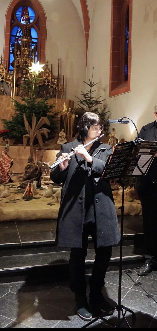 Person in schwarzem Mantel spielt Querflöte in einer Kirche vor Weihnachten, mit Weihnachtsbäumen und Krippenszene im Hintergrund, Notenständer vor ihr