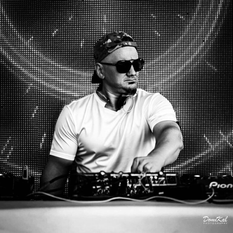 Schwarz-weiß-Foto eines DJs mit Sonnenbrille und Rückwärtskappe, der hinter seinem Mischpult steht und eine Hand darauf legt, vor einem Bildschirm mit Lichtpunkten.