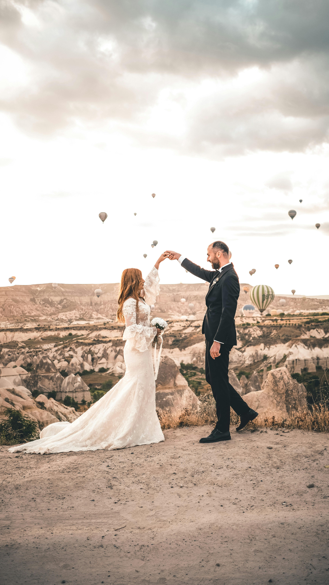 Braut in weißem Spitzenkleid und Bräutigam im schwarzen Anzug tanzen auf einer felsigen Anhöhe mit vielen bunten Heißluftballons am Himmel im Hintergrund an einem bewölkten Tag.