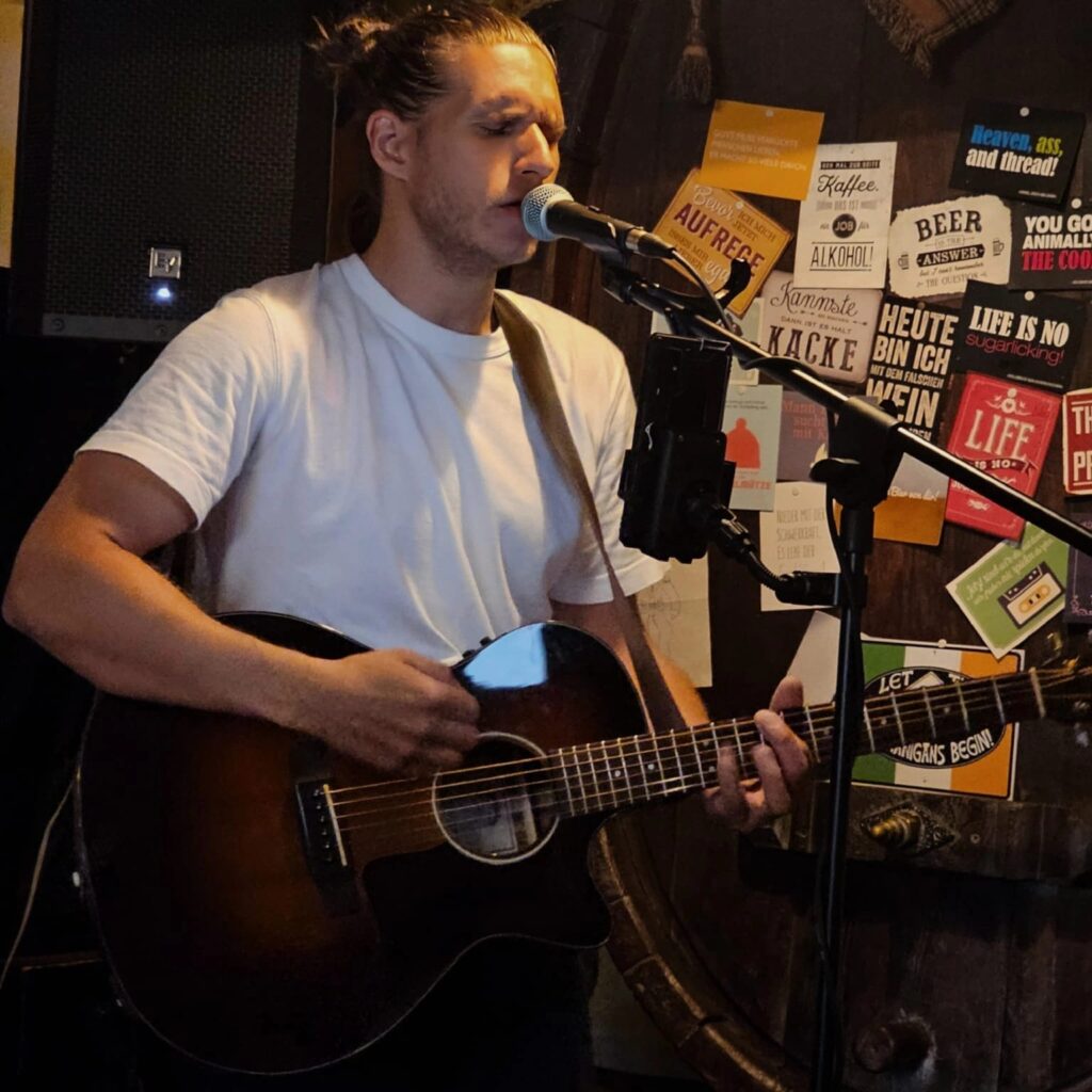 Mann mit weißem T-Shirt, der vor einem Mikrofon singt und auf einer dunklen Akustikgitarre spielt, vor einer Wand mit verschiedenen bunten Aufklebern und Sprüchen im Hintergrund