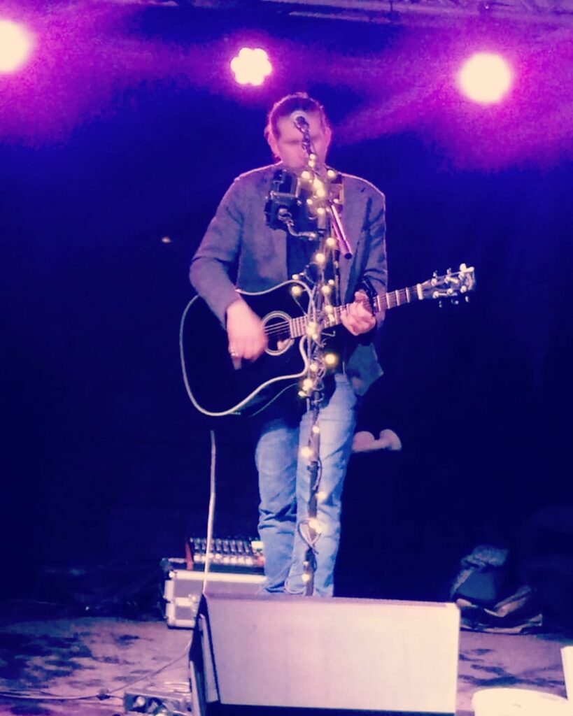 Ein Musiker spielt auf einer Bühne eine Akustikgitarre, ein Mikrofonständer mit Lichterkette steht davor, und im Hintergrund leuchten violette Bühnenlichter.