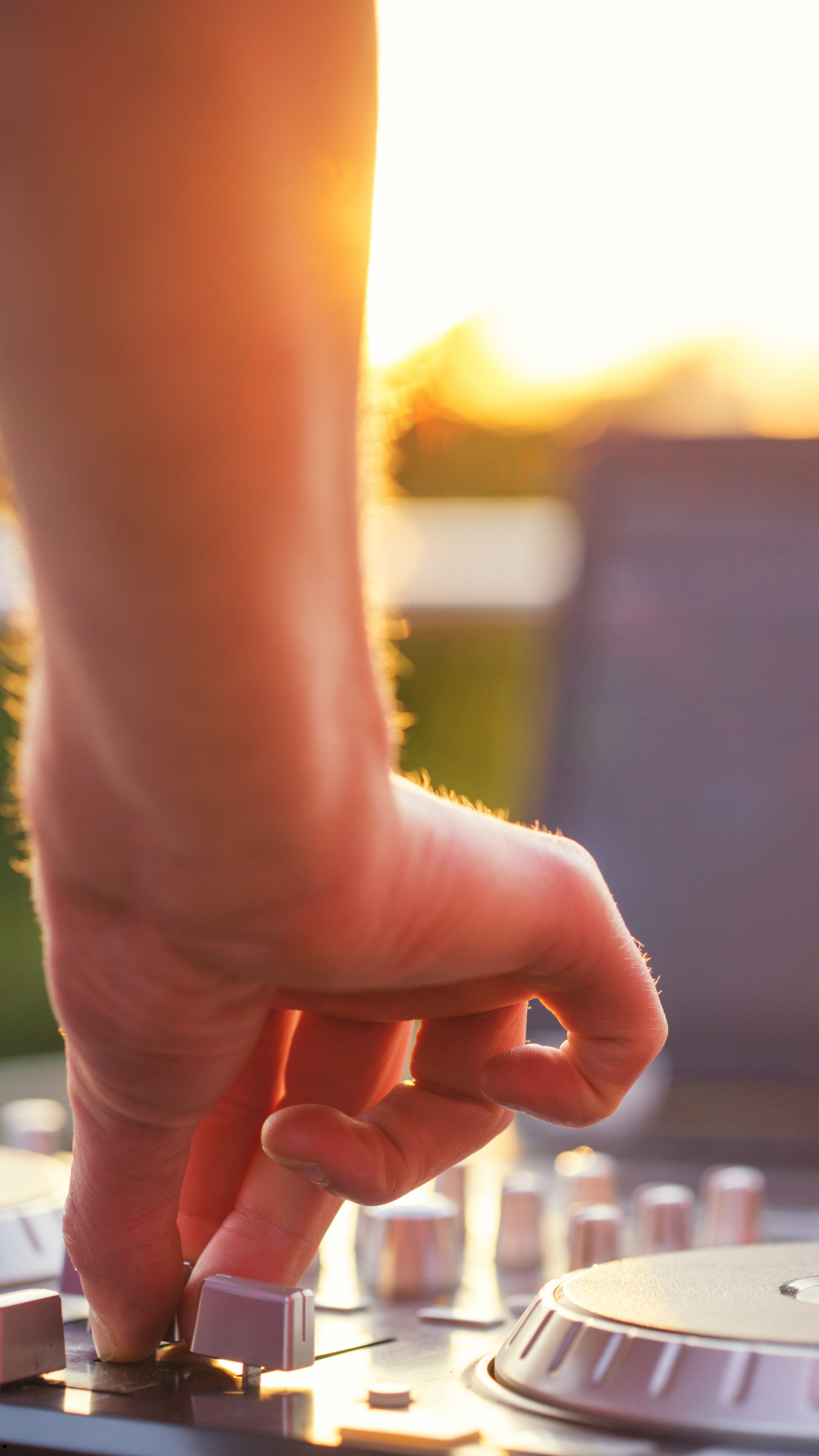 Nahaufnahme einer Hand, die an einem Regler eines DJ-Mischpults bei Sonnenuntergang dreht, mit unscharfem Hintergrund und warmem Licht