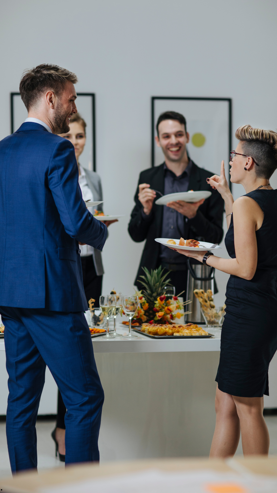 Vier beruflich gekleidete Personen stehen um einen Buffet-Tisch mit Getränken und Snacks und unterhalten sich bei einem Event oder Empfang.