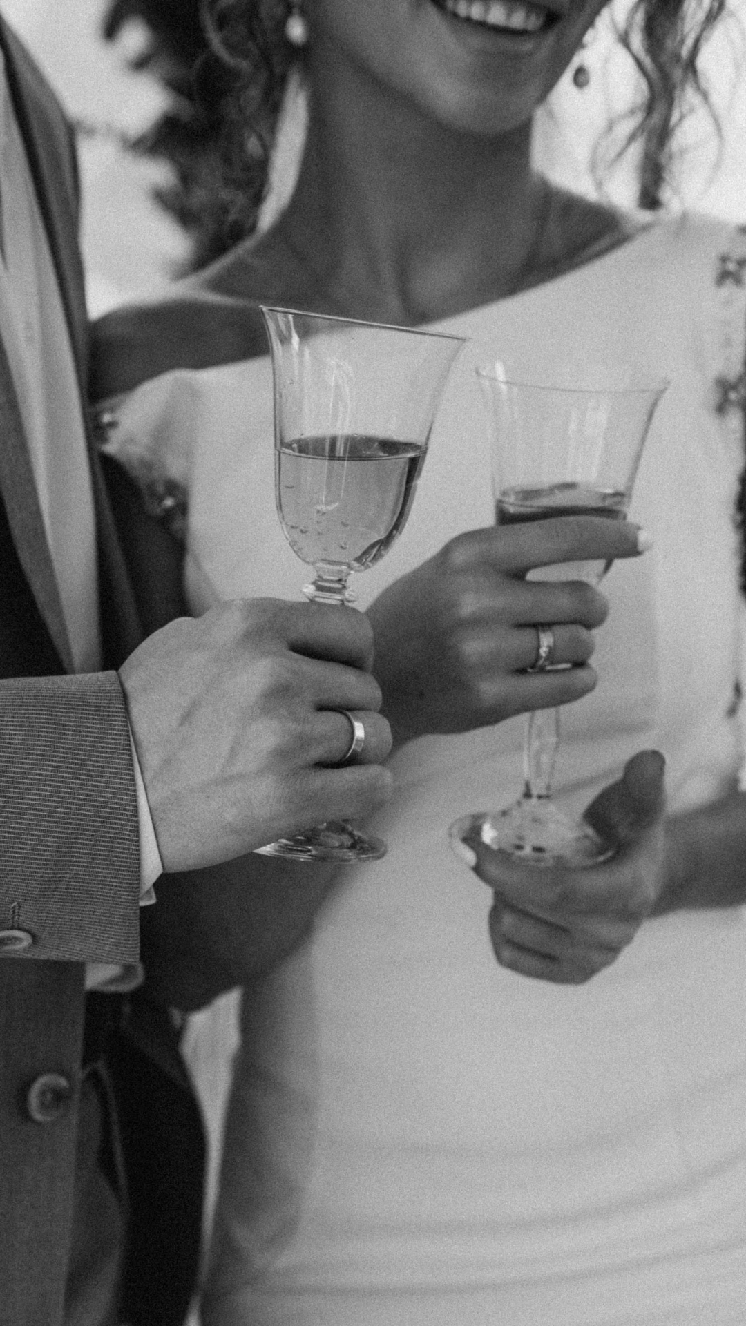 Schwarz-weiß-Foto eines Paares, das mit Sektgläsern anstößt, die Hände zeigen Eheringe, die Frau trägt ein weißes Kleid und lächelt.
