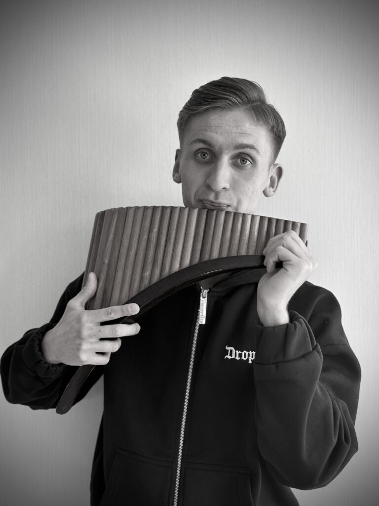 Schwarz-weiß-Porträt eines jungen Mannes mit kurz zurückgekämmtem Haar, der eine Panflöte spielt und eine Jacke mit Reißverschluss trägt.