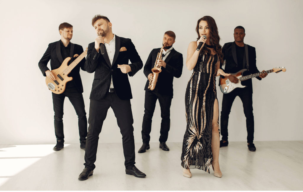 Band mit zwei Sängern, einer Frau im schwarzen und goldenen Abendkleid und einem Mann im schwarzen Anzug, sowie drei Musikern mit Bass, Saxophon und E-Gitarre vor weißem Hintergrund. Jetzt Hochzeitsband buchen!