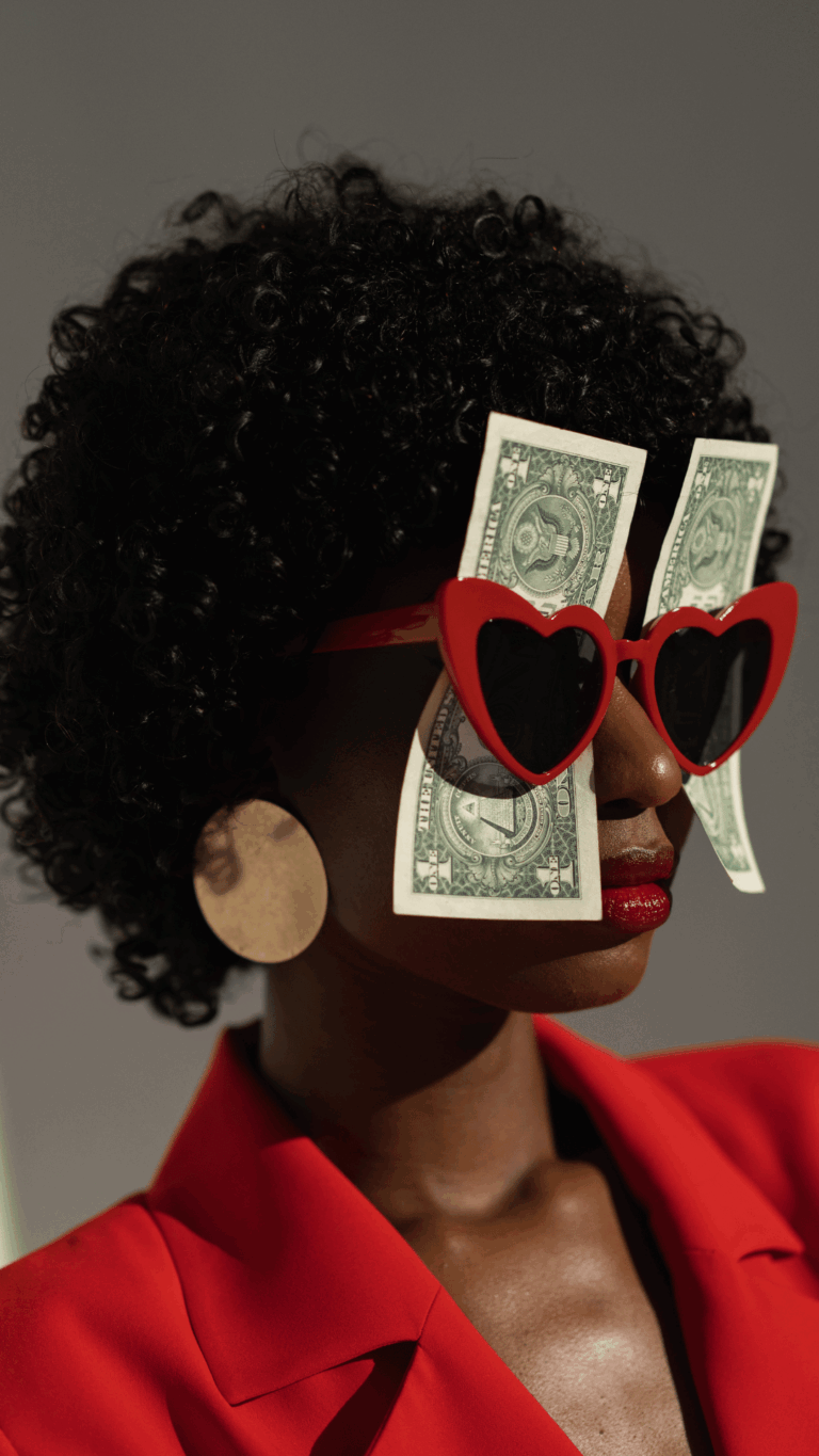 Porträt einer Frau mit lockigem schwarzen Haar, die rote Herzförmige Sonnenbrille trägt, in die zwei Ein-Dollar-Scheine gesteckt sind, roter Lippenstift und roter Blazer, große runde goldene Ohrringe, vor grauem Hintergrund.