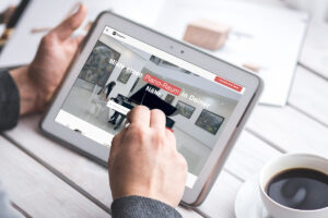 Person hält ein Tablet, auf dem eine Webseite zum Mieten eines Piano-Raums gezeigt wird, im Hintergrund ist ein Becher mit Kaffee auf einem weißen Tisch zu sehen