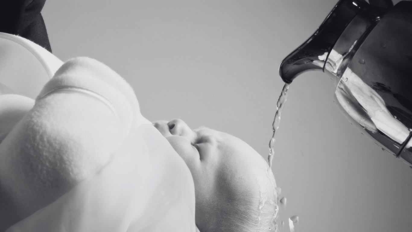 Schwarz-weiß-Foto eines Babys, dem aus einem Wasserkessel Wasser über den Kopf gegossen wird, mit geschlossenen Augen und entspanntem Gesichtsausdruck. Titelbild für den Artikel Musik für die Taufe.