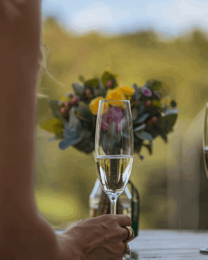 Sektempfang im Frühling – Jahreszeit für eure Hochzeit. Hand mit Ringen hält ein Sektglas vor einem unscharfen Hintergrund mit einem Blumenstrauß mit gelben und lila Blüten.