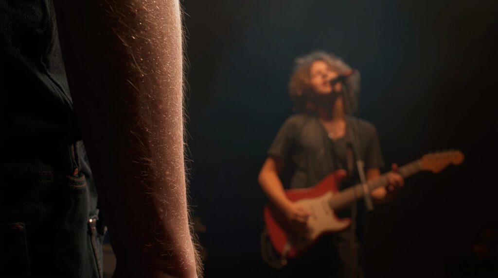 Nahaufnahme eines beleuchteten Armen mit Körperbehaarung im Vordergrund, im unscharfen Hintergrund ein Musiker mit Locken, der eine E-Gitarre spielt und vor einem Mikrofon singt. Musik und Erinnerungen schaffen Gänsehaut.