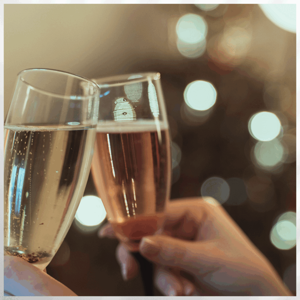 Zwei angestoßene Sektgläser, eins mit hellem und eins mit roséfarbenem Sekt, vor einem unscharfen Hintergrund mit Lichtern.