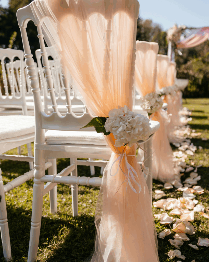 Hochzeit im Freien im Sommer: Jahreszeit für eure Hochzeit. Reihe weißer Stühle im Freien, dekoriert mit zartem, peachfarbenem Stoff und weißen Blumen, auf einem Rasen mit verstreuten Blütenblättern, vermutlich für eine Hochzeit oder festliche Zeremonie.