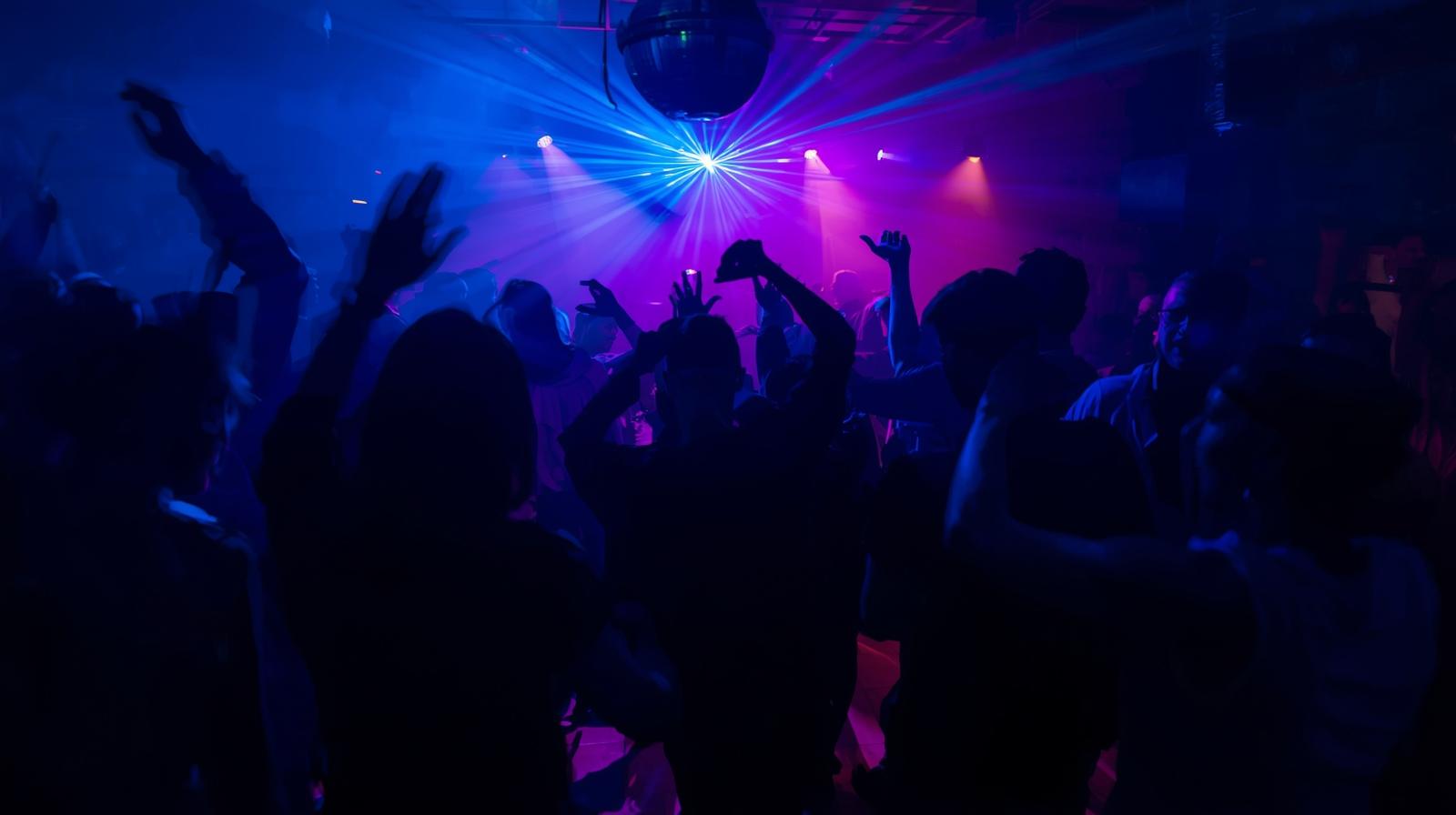 Dunkle Silhouetten von Menschen, die in einem Club mit blauer und violetter Beleuchtung und einer Diskokugel tanzen und ihre Hände in die Luft heben. Wer wohl Musik macht? DJ oder Band?