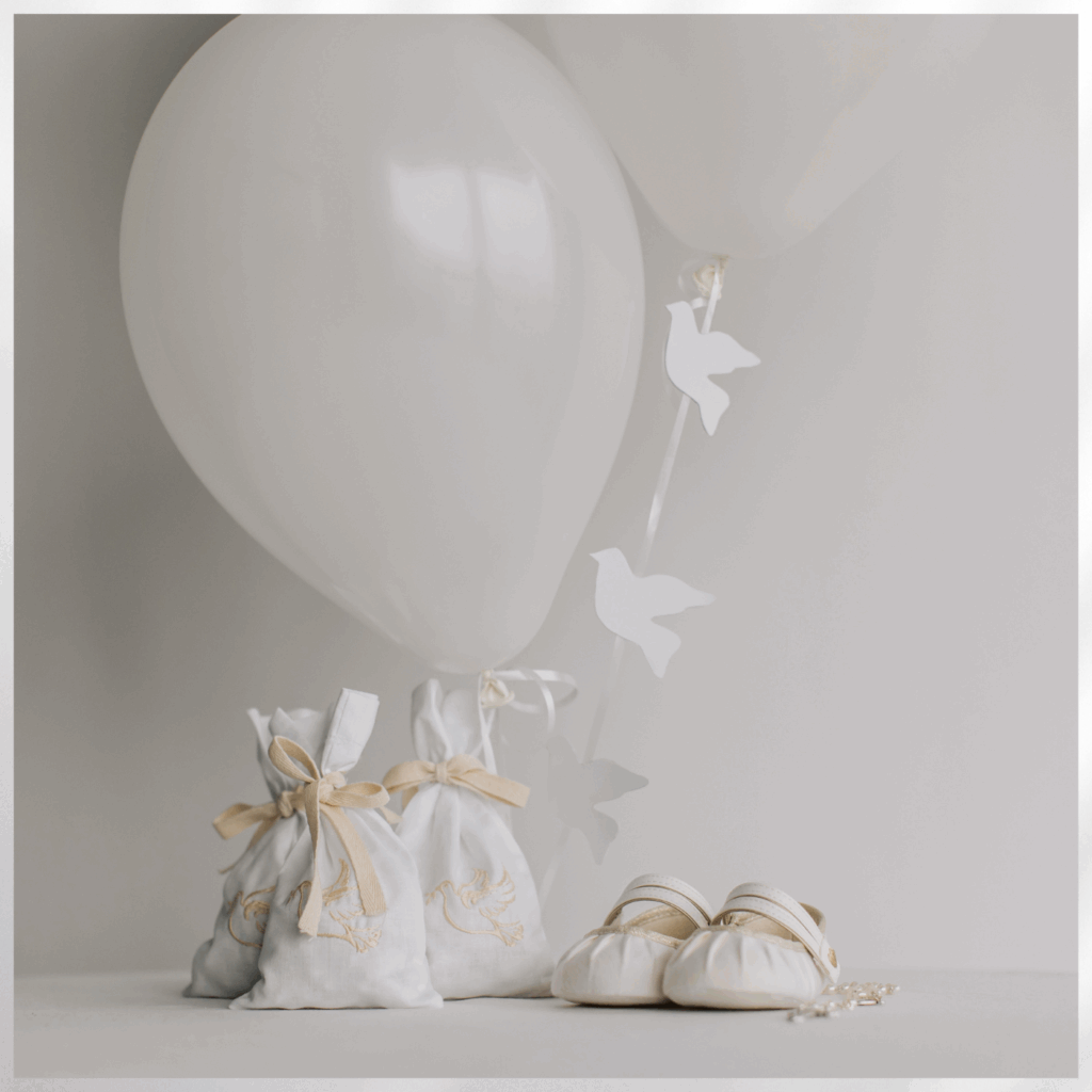Drei weiße Stoffbeutel mit goldenen Schleifen und Stickereien neben einem Paar weißer Babyschuhe, im Hintergrund weiße Luftballons und Papiervögel an einer Schnur vor hellgrauer Wand.