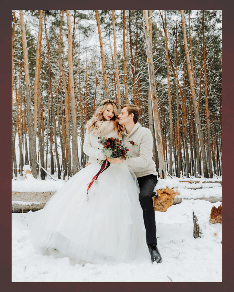 Winterhochzeit im Schnee – Jahreszeit für eure Hochzeit. Brautpaar im Winterwald, die Braut trägt ein weißes Hochzeitskleid mit Strickjacke und Schal, hält einen Blumenstrauß, der Bräutigam trägt einen hellen Strickpullover und schwarze Hose, sie sitzen auf einem Baumstamm im Schnee und schauen sich liebevoll an.