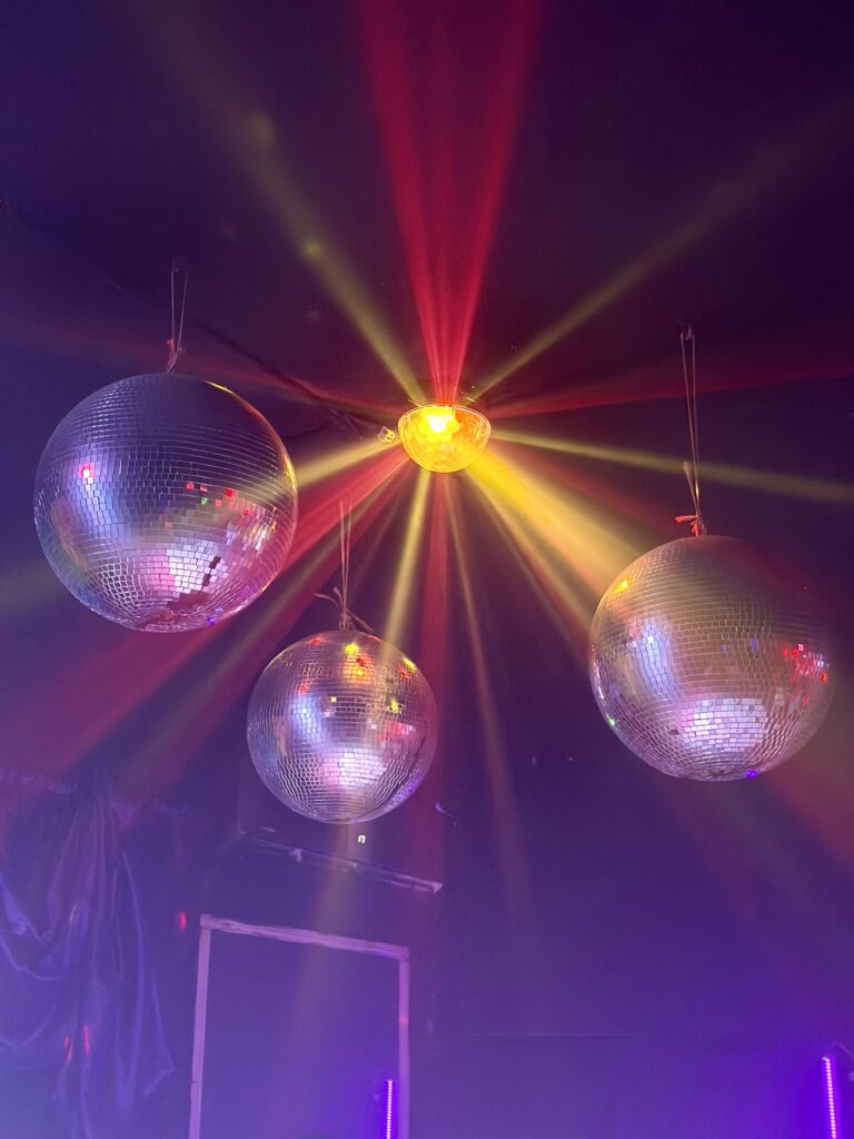 Bunte Bühnenlichter schaffen Stimmung auf einer Veranstaltung. Drei spiegelnde Discokugeln hängen unter einer Decke, von der eine Lichtquelle bunte Strahlen ausstrahlt und die Kugeln reflektieren das Licht in einem dunklen Raum mit farbigen Beleuchtungseffekten.