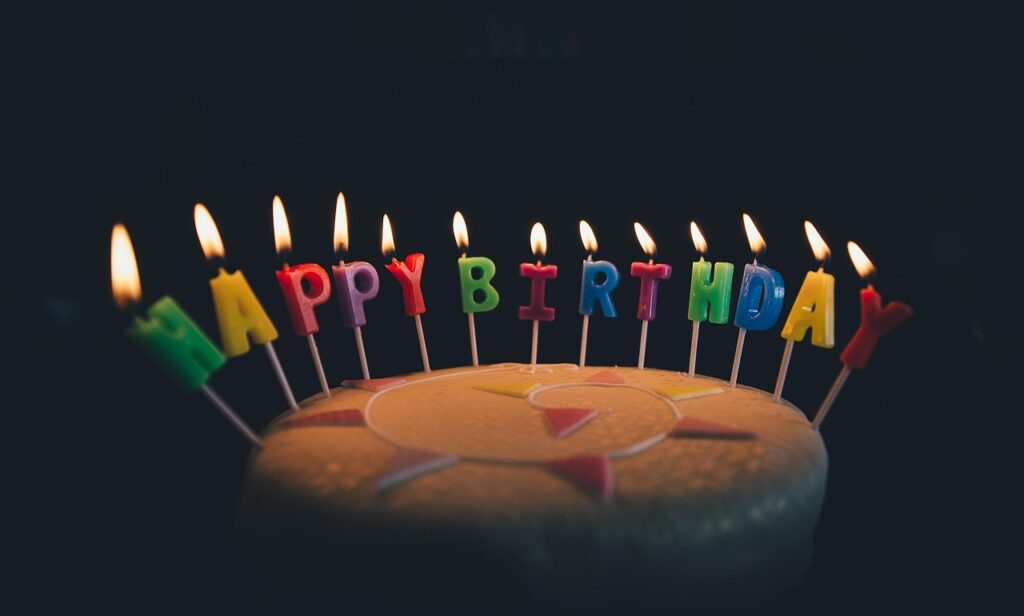 Geburtstagskuchen mit brennenden, bunten Kerzen, die die Buchstaben HAPPY BIRTHDAY bilden, dunkler Hintergrund