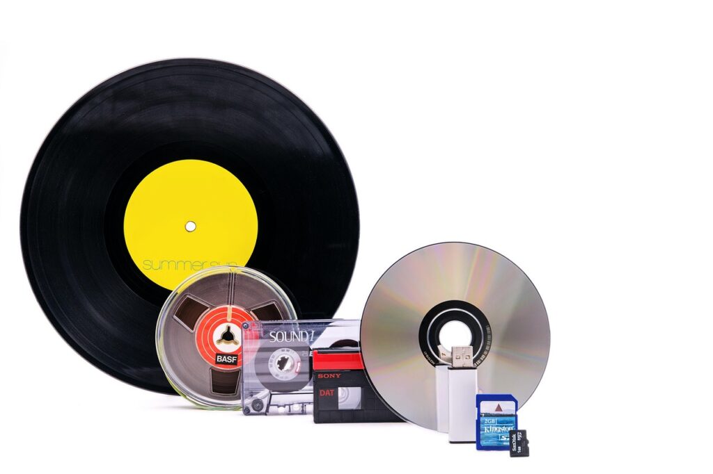 Verschiedene Speichermedien der Musikgeschichte in einer Reihe, darunter eine schwarze Schallplatte mit gelbem Etikett, eine Spulentonbandkassette, eine Audio-Kassette, eine DAT-Kassette, eine CD, ein USB-Stick, eine SD-Karte und eine Mikro-SD-Karte.