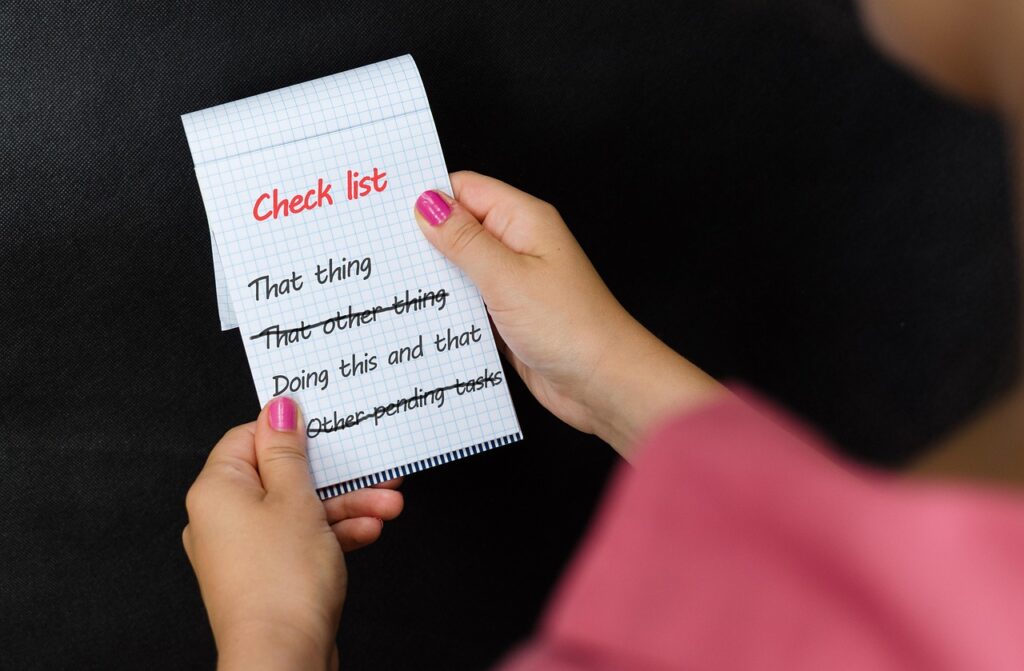 Frau mit pinkem Oberteil hält einen karierten Notizblock mit der Überschrift Check list, darauf stehen verschiedene Aufgaben, von denen einige durchgestrichen sind. Gastgeber:in prüft eine Checkliste für Technik & Aufbau bei einem Event.