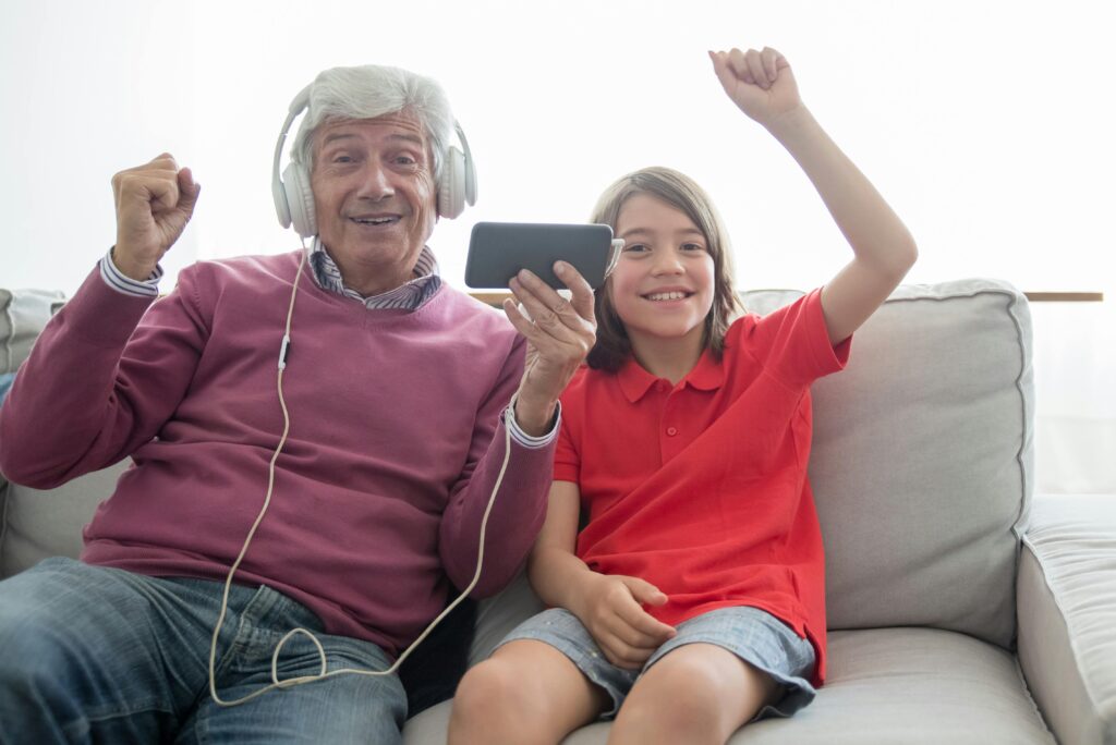 Ein älterer Mann mit weißen Haaren trägt Kopfhörer und hält ein Smartphone, sitzt gemeinsam mit einem lachenden Jungen in rotem Shirt und grauen Shorts auf dem Sofa, beide haben eine Hand zum Jubeln erhoben.