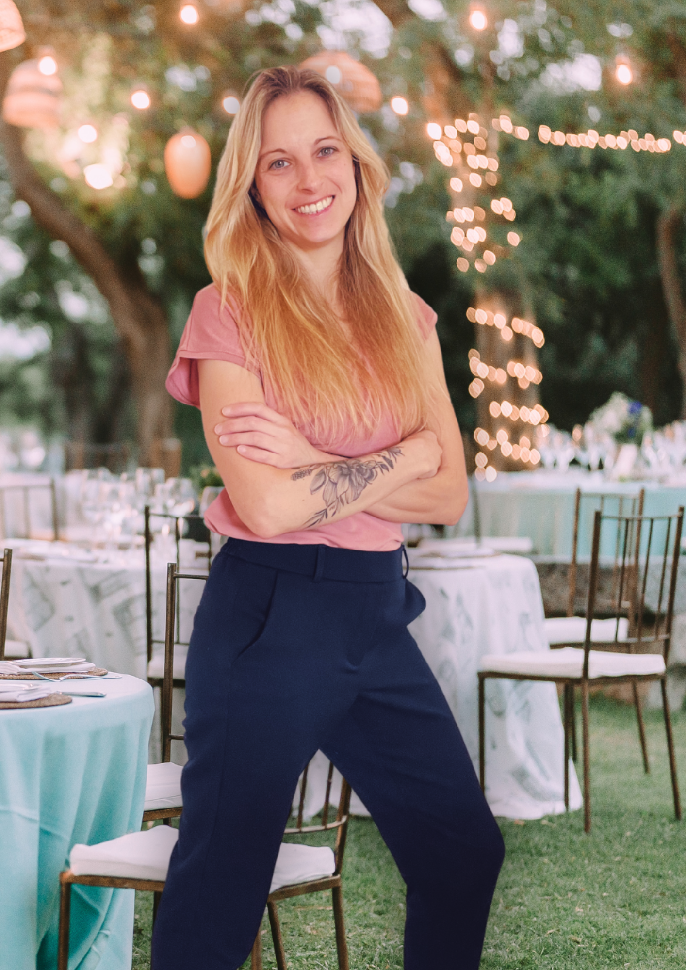 Hochzeitsplanerin Bayern: Junge Frau mit langen blonden Haaren, rosa T-Shirt und blauer Hose steht mit verschränkten Armen lächelnd in einem Garten mit festlich gedeckten Tischen und Lichterketten im Hintergrund.