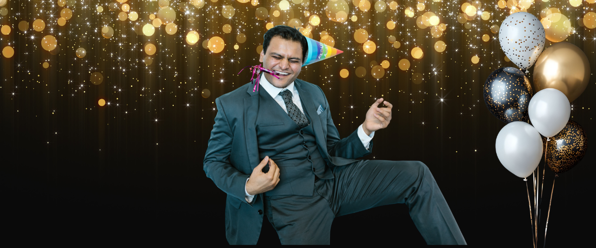 Musik für die Firmenfeier: Mann in Anzug mit Partyhütchen und Luftschlange im Mund, der freudig tanzt vor einem festlichen Hintergrund mit goldenen Lichtern und einem Strauß aus schwarzen, weißen und goldenen Luftballons.